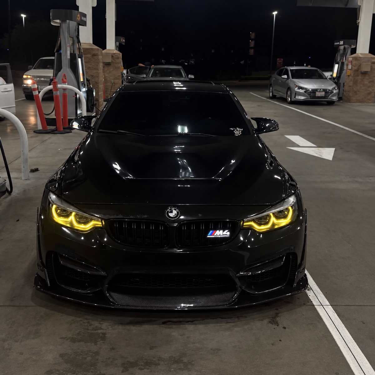 Design visualization of a solid-white illuminated Velor Works M4 Competition emblem on a black BMW at a gas station; digitally edited image, not a fitment certification